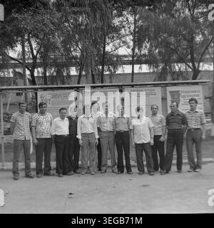 Sloviansk, 1981, AIZ (Armature-Insulator Plant). A group of eleven male factory workers poses for a portrait outdoors in the courtyard, standing in front of large informational boards displaying Soviet socialist obligations and labor safety metrics. Tall trees and industrial buildings provide the backdrop for this proud collective of employees. A historical black and white archive photograph capturing the authentic atmosphere of the era, the camaraderie of the working class, and the peaceful Donbas. Stock Photo