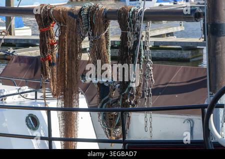 Fishing nets and ropes hang on a rail at a harbor where boats are docked. The scene shows different types of fishing gear in bright sunlight on a clea Stock Photo