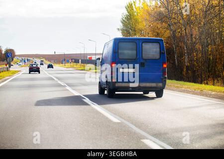 Rear view of blue delivery van moving along asphalt highway, cars visible in distance, vibrant autumn trees alongside road. Clear sky overhead, utilit Stock Photo