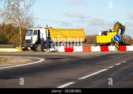 Daytime view of road construction project featuring large yellow dump truck, excavator, and two road workers standing next to a line of red and white Stock Photo