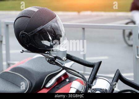 Closeup view of black motorcycle helmet visor and shell, covered in water droplets from recent rain, resting on chrome handlebars of contemporary moto Stock Photo