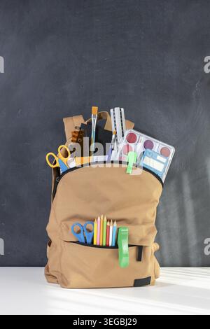 School backpack overflowing with Stationery Supplies on blackboard with copy space, ready for back to school season Stock Photo