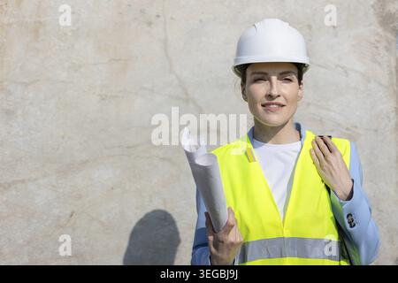 Portrait of a confident female architect wearing a protective vest, smiling and holding blueprints at a construction site. Copy space Stock Photo
