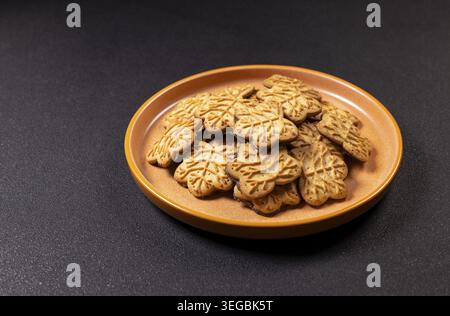 Maple leaf shaped cookies, glazed with chocolate filling, arranged on an orange ceramic plate, offer a sweet snack on a textured dark background for f Stock Photo
