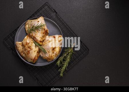 Raw chicken legs are marinated on a gray plate with fresh rosemary and various spices, placed on a black rack against a dark background and prepared f Stock Photo