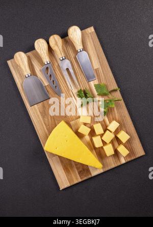 Wooden board with a cheese wedge, small cheese cubes and a set of cheese knives made of stainless steel and wood with fresh parsley, prepared for serv Stock Photo