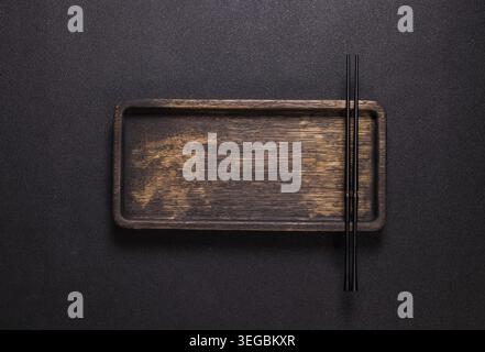 Black chopsticks lie on a rectangular wooden board or serving tray, which is positioned against a dark, textured background and creates a minimalistic Stock Photo