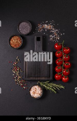 Dark wooden cutting board with cherry tomatoes on the vine, fresh rosemary sprig, coarse salts and mixed peppercorns, arranged with spices on a black Stock Photo