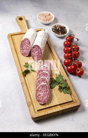 Dried salami, whole and sliced, on a wooden cutting board with fresh parsley, cherry tomatoes, peppercorns and pink salt to make an appealing starter Stock Photo