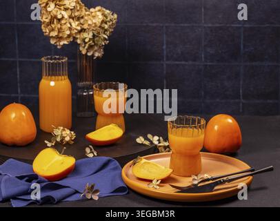 Two glasses and a carafe of persimmon juice served with slices of fresh persimmon on a dark table with dried flowers make for a cozy fall scene Stock Photo