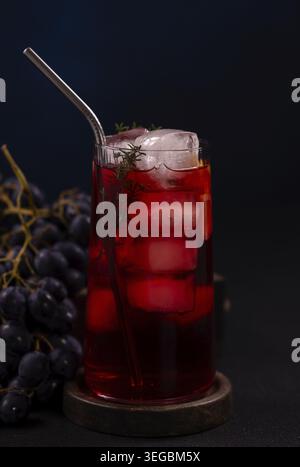 Tall glass of chilled red grape juice with ice, thyme branch and metal straw on a trivet, studio shot on dark background with blurry grapes and copy s Stock Photo