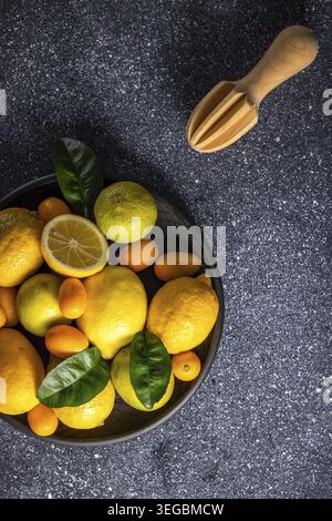 Vivid citrus fruits such as lemons, limes and kumquats arranged in a black bowl next to a wooden reamer and create a refreshing scene on a dark stone Stock Photo
