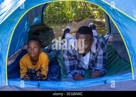 African American father in plaid, son in yellow lying on pads in blue tent holding binoculars Stock Photo