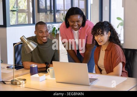 Diverse coworkers collaborating around laptop at office desk, leaning over screen with brass lamp Stock Photo