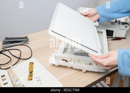 Hands carefully disassembling a flatbed scanner on a wooden desk, showing internal parts, cables and tools during repair and maintenance in a home off Stock Photo