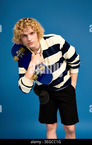 Blonde, curly haired student showcases style while holding a skateboard in a lively studio. Stock Photo