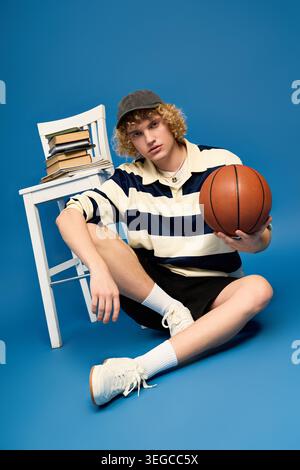 Blonde, curly haired student showcases fashion while sitting with a basketball in studio. Stock Photo