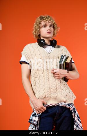 Blonde curly haired young man showcases his trendy outfit while holding books and headphones. Stock Photo