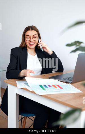 Beautiful woman talking on phone and reviewing documents at office desk with laptop. Business. Office work. Female professional analyzing papers. Beau Stock Photo