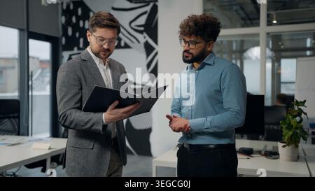 Two multiracial business men talking looking in folder with papers in office diverse businessmen Caucasian leader reading report contract read data Stock Photo