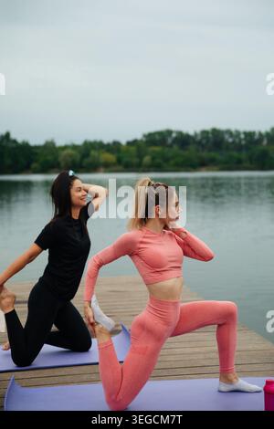 Two women are practicing yoga on a wooden dock by a calm lake. They are engaged in different poses, promoting flexibility and harmony in their fitness Stock Photo