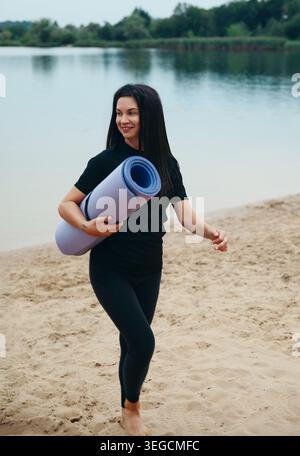 A woman walks on the sandy beach, holding a yoga mat, ready for a fitness session by the water. She is smiling, embracing wellness and meditation. Stock Photo