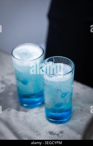 Two tall clear glasses filled with bright blue chilled beverage, ice cubes and white foam stand on a gray stone surface. Stock Photo