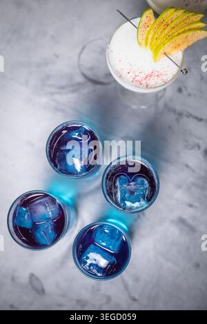 Overhead shot of four blue alcoholic drinks with ice cubes and one apple cocktail on a concrete background. Stock Photo