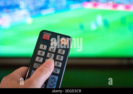 Watching football match at home, close up on hand holding remote with defocused tv screen of football match at the background. Stock Photo