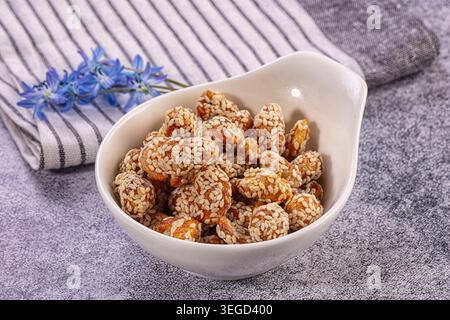White bowl filled with delicious sesame coated nuts, likely caramelized peanuts, rests on gray textured surface. Crisp white golden brown snack presen Stock Photo