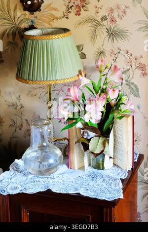 Cozy vintage-style bedside table featuring pastel pink floral arrangement and an open book, lampshade, carafe against floral wallpaper. Stock Photo