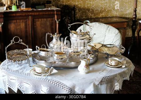 Ornate Victorian-style silver tea service and fine china teacups on a white lace tablecloth inside a stately home dining room. Stock Photo