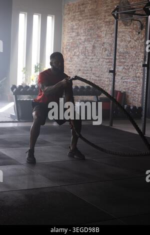 African American man performing battle rope waves in industrial gym wearing red shirt, black shorts Stock Photo