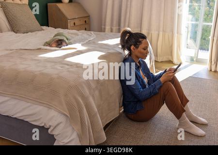 Mid-adult woman in denim shirt and socks sitting by bed using smartphone watching baby, copy space Stock Photo
