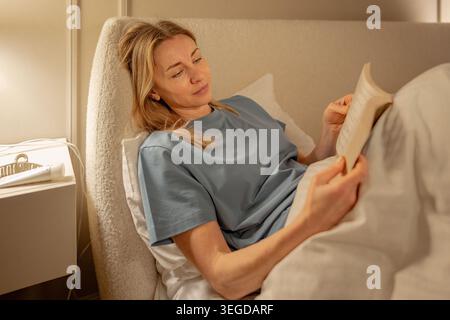 Woman lying in bed, reading book during quiet evening at home. Calm bedtime routine, relaxation, slow living, digital detox concept. Peaceful female e Stock Photo