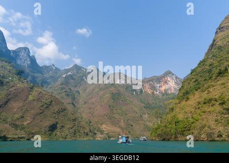 Serene lake surrounded by towering green mountains. Small boats drift on calm, turquoise waters. Clear blue sky with soft white clouds above. Rugged c Stock Photo