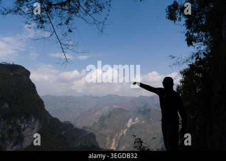 Silhouette of person pointing toward vast mountain range. Deep valleys stretch beneath layered peaks. Clouds drift above rugged, forested cliffs. Dapp Stock Photo