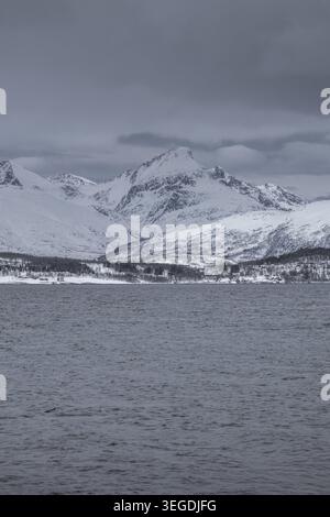 View of snow-covered mountains rising above the cold waters of a fjord under a dark overcast sky Tromso, Troms, Norway. Stock Photo