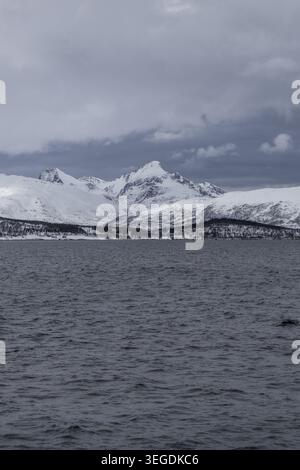 View of majestic snow-capped mountains rising above the dark waters of a fjord under an overcast sky in Tromso, Troms, Norway. Stock Photo