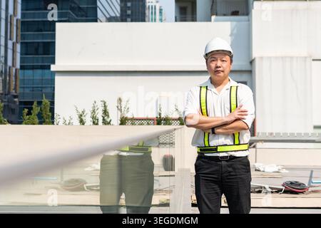Confident construction supervisor standing with crossed arms while overseeing operations at modern construction site. Stock Photo