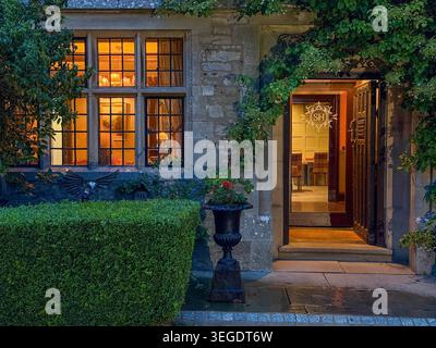 View of a historic stone manor house entrance with warm interior lighting through leaded windows, an open doorway, and lush garden greenery Hawes, Eng Stock Photo
