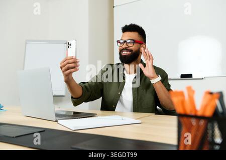 Young handsome man smiles and waves during a video call in a modern office space. Stock Photo