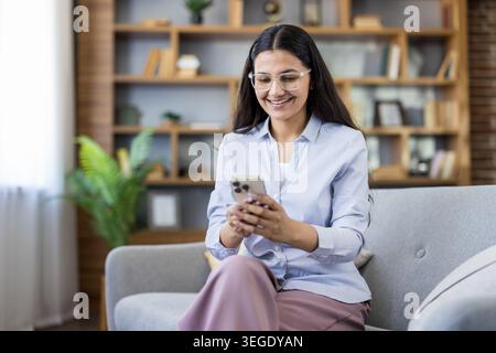 Woman wearing glasses sitting on a comfortable sofa in a modern living room, smiling happily while texting on her mobile phone, connecting and communicating online with technology Stock Photo