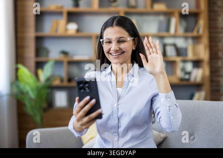 Young smiling woman with glasses waving during a smartphone video call at home, enjoying casual conversation with friends or family via online app, warm and cheerful connection Stock Photo