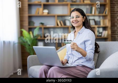 Young indian woman sitting on a sofa, holding a credit card and using a laptop for online shopping, smiling at the camera, depicting e-commerce and secure banking from home Stock Photo