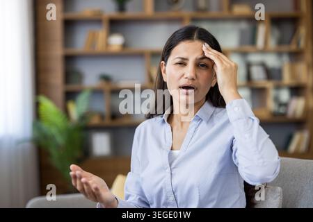 Young adult woman feeling overwhelmed, stressed, or experiencing a headache, placing a hand on her temple while sitting indoors in a home or office environment Stock Photo
