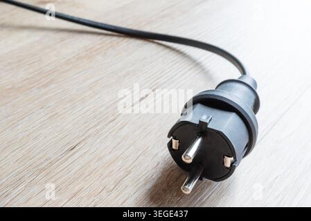 close-up of black Schuko safety power plug with its cable lying on a light-colored wooden floor Stock Photo