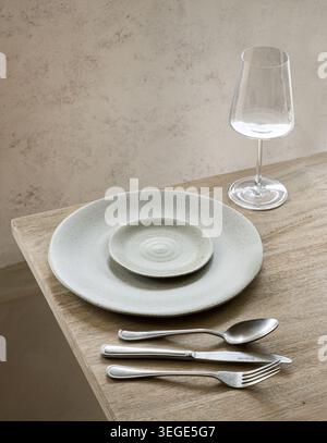 A rustic table setting featuring speckled ceramic plates, silver cutlery, and an empty wine glass on a wooden surface against a textured background. Stock Photo