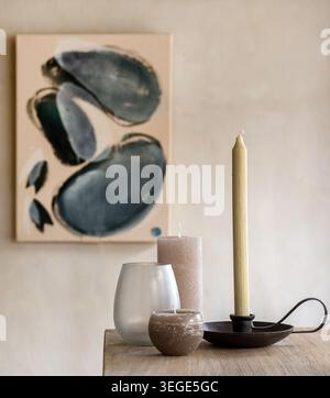 A collection of various decorative candles, a glass holder, and a metal candlestick holder on a wooden table with an abstract canvas painting in the b Stock Photo