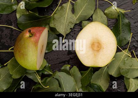 a ripe juicy sliced pear lies on the table with the green foliage of the pear, a large number of green leaves of the pear tree and a ripe covered pear Stock Photo